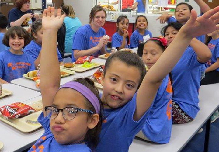 Two children smiling and posing in a cafeteria with classmates in the background.