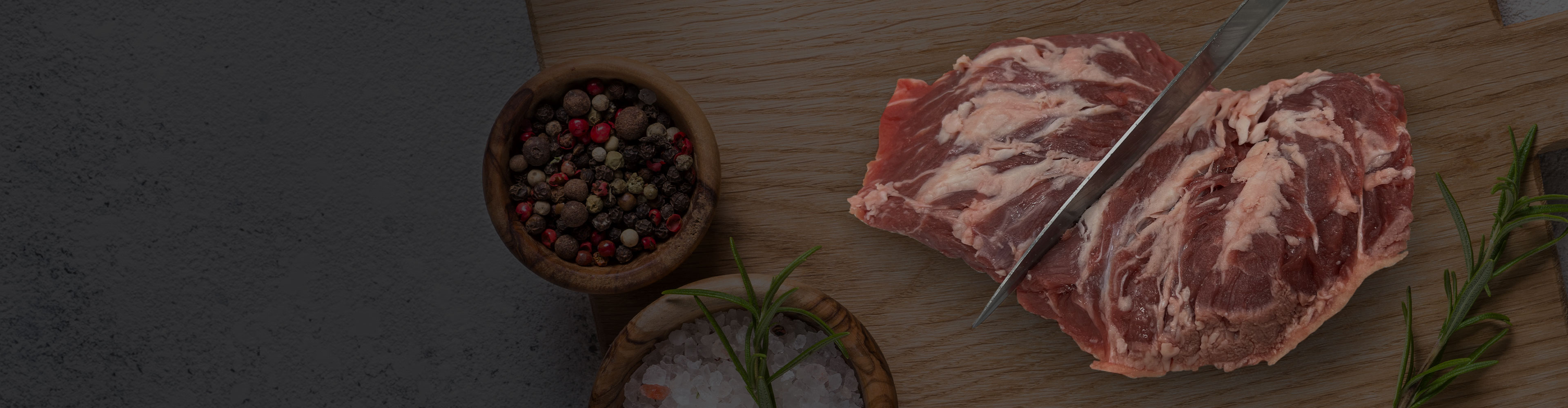 Raw beef steak being sliced on a wooden cutting board with peppercorns, coarse salt, and rosemary nearby.