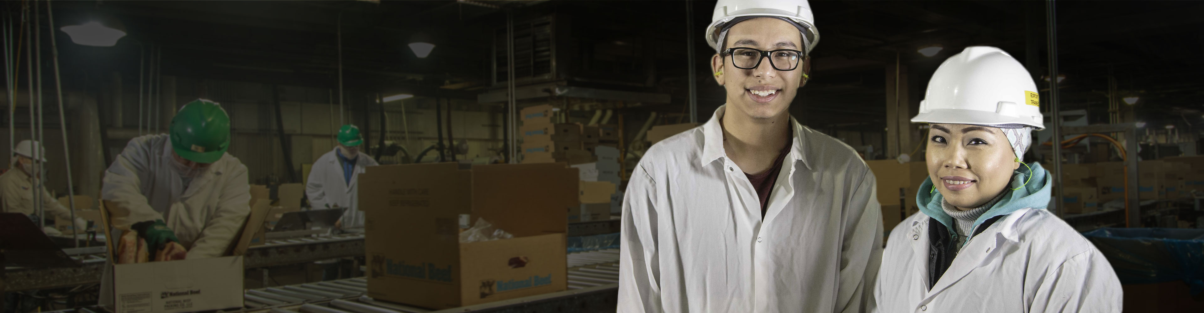 Two workers in protective gear and white hard hats standing in a processing facility with coworkers packing boxes in the blurred background.