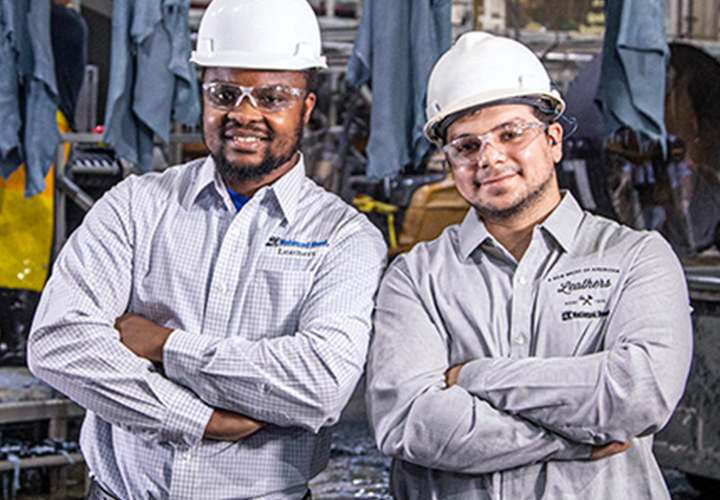 Two employees in protective gear smiling while standing inside a leathers facility.