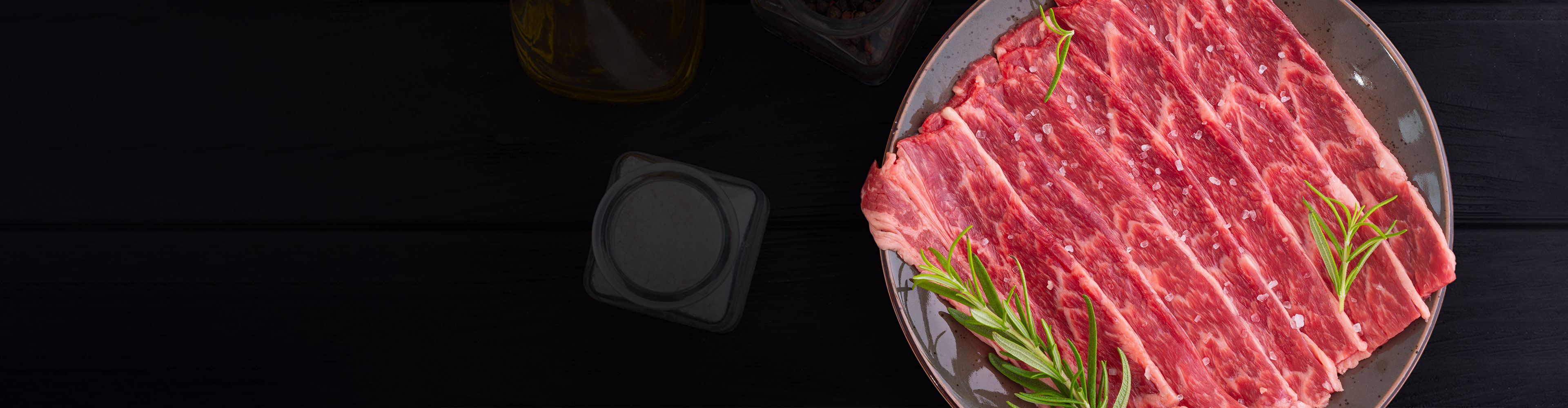 生の牛肉のスライスがローズマリーと調味料とともに暗い背景に皿に並べられています。