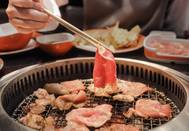 Thin slice of raw beef held over a grill with additional pieces cooking below and blurred dishes in the background.