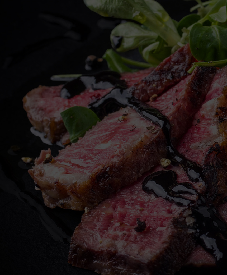 Slices of seared beef drizzled with sauce and served with greens on a dark plate.