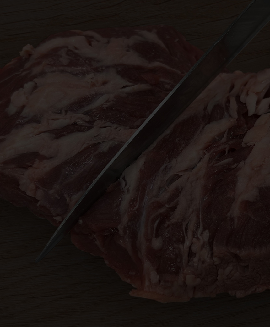 Raw beef steak being sliced on a wooden cutting board with peppercorns, coarse salt, and rosemary nearby.