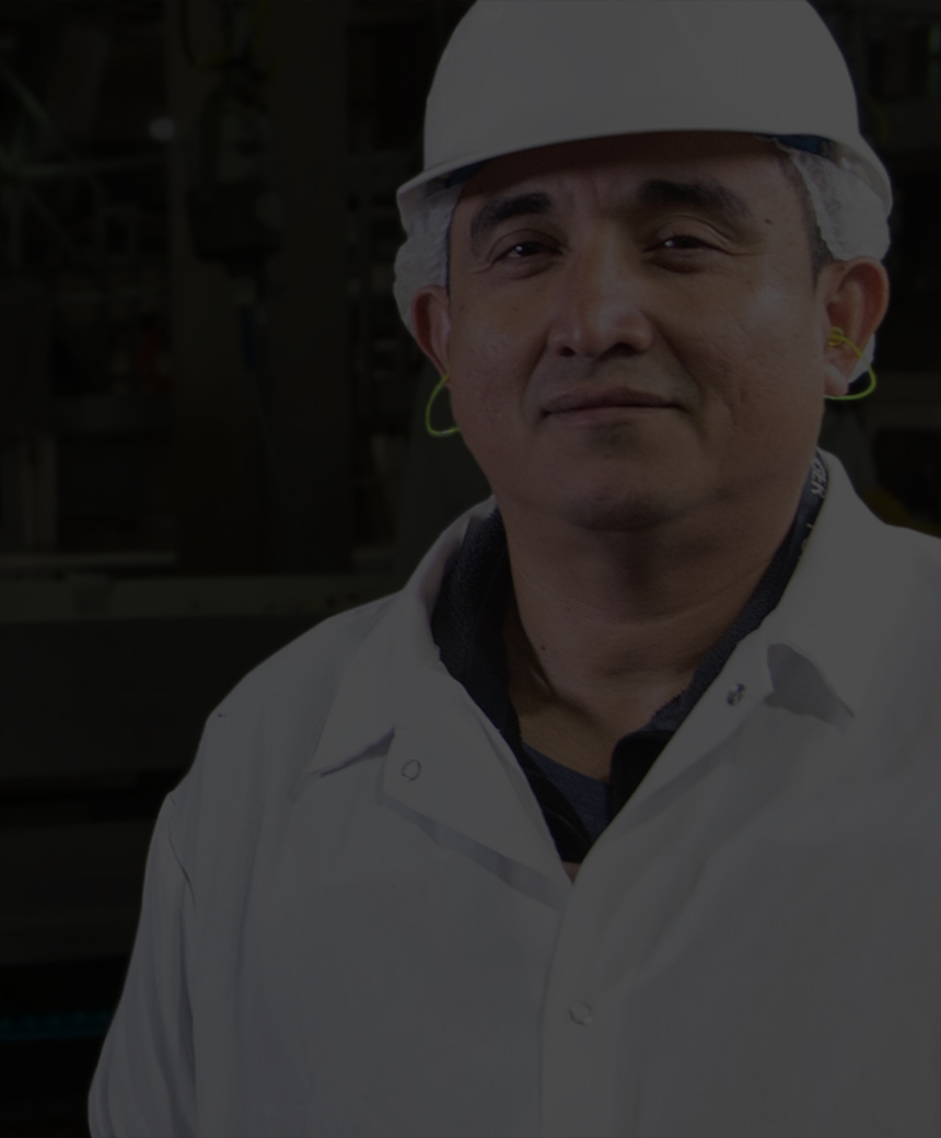Worker in a white coat and hard hat standing in front of a production line inside a processing facility.