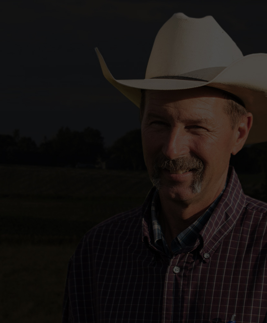 Smiling rancher in a cowboy hat standing in an open field with grazing cattle and a distant farm on the horizon.