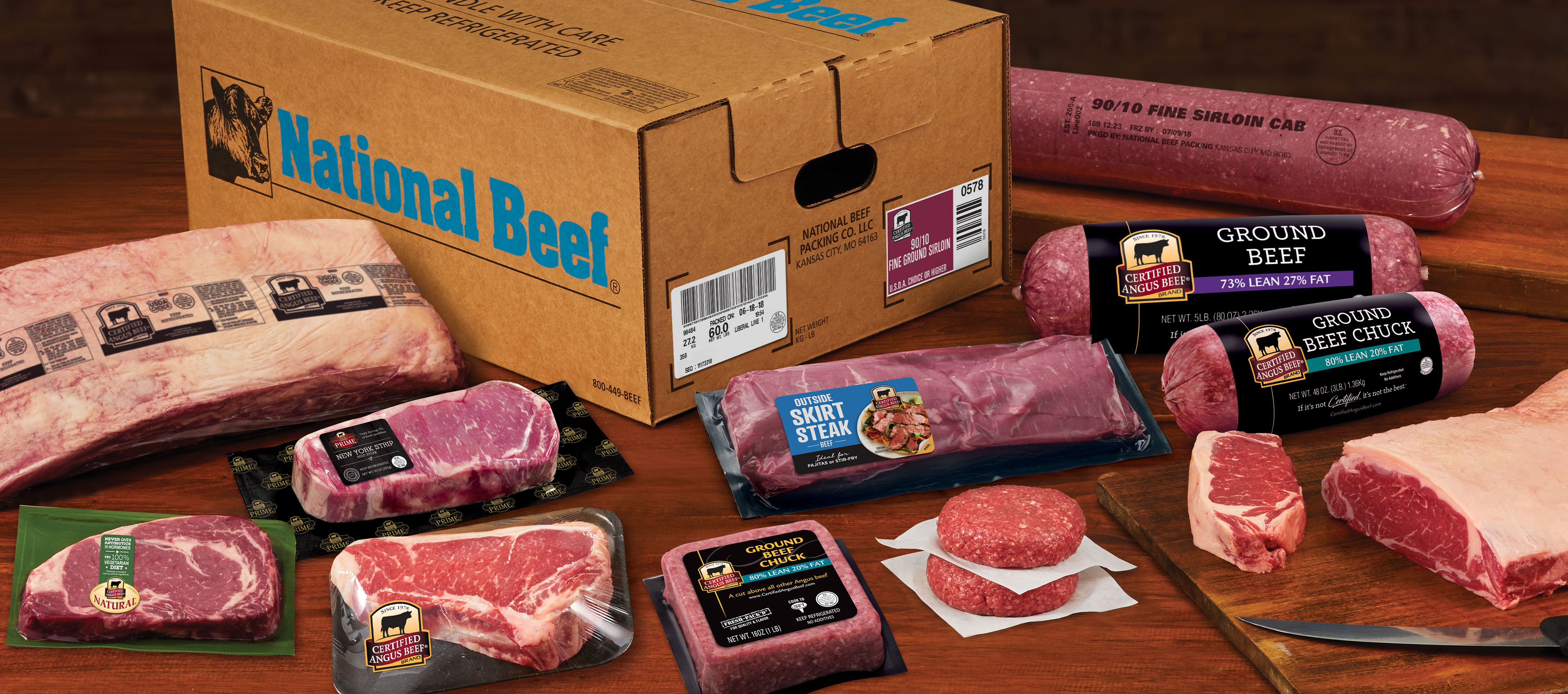 National Beef shipping box surrounded by assorted Certified Angus Beef primals, steaks, ground beef chubs, and patties on a wooden surface