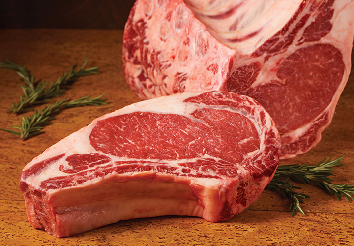 Raw bone-in rib roast and a ribeye steak displayed on a wooden board with rosemary sprigs