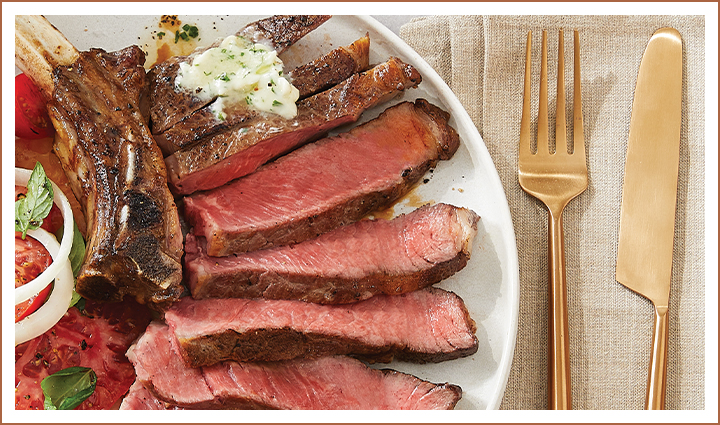 Cooked sliced steak with bone on plate beside tomatoes, onions, and gold flatware