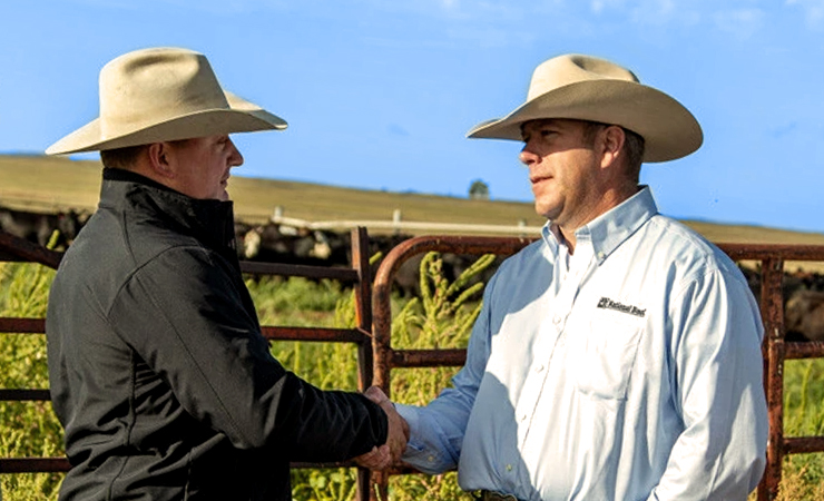 Two cattlemen in hats shaking hands by a corral fence