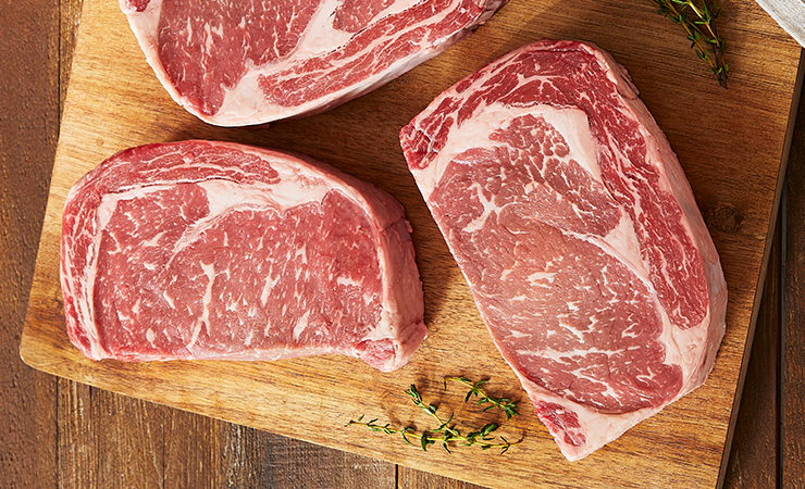Three raw ribeye steaks on a wooden cutting board with herbs and a small dish of compound butter