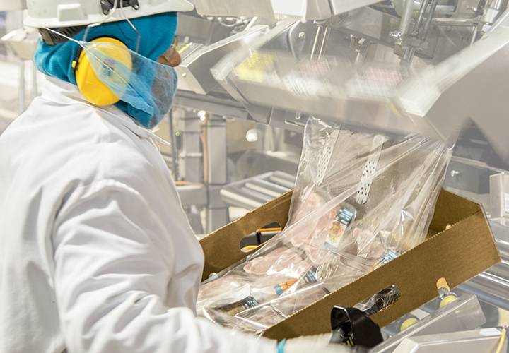 Worker in protective gear handling packaged meat products on a processing line.