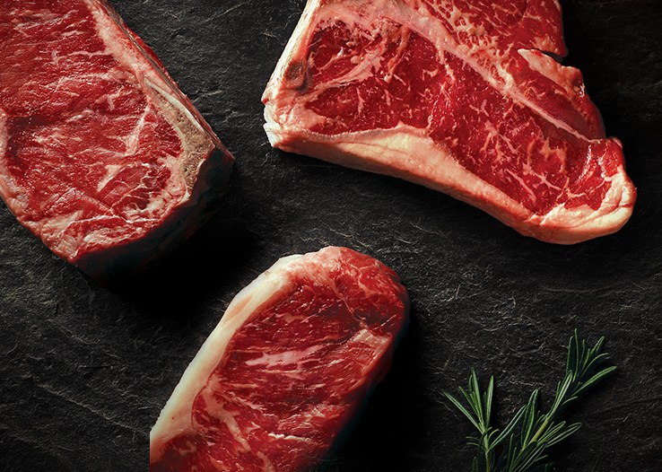 Raw steaks arranged on a dark textured surface with a sprig of rosemary.