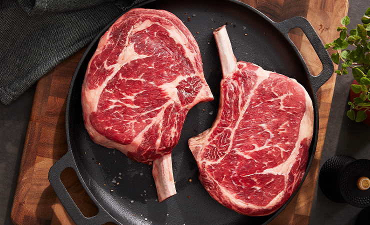 Two raw bone-in ribeye steaks in a round cast-iron pan on a wooden board with seasonings nearby