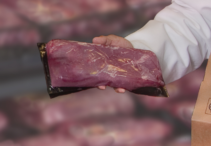 Person in a white coat holding a vacuum-sealed beef cut next to a cardboard shipping box.