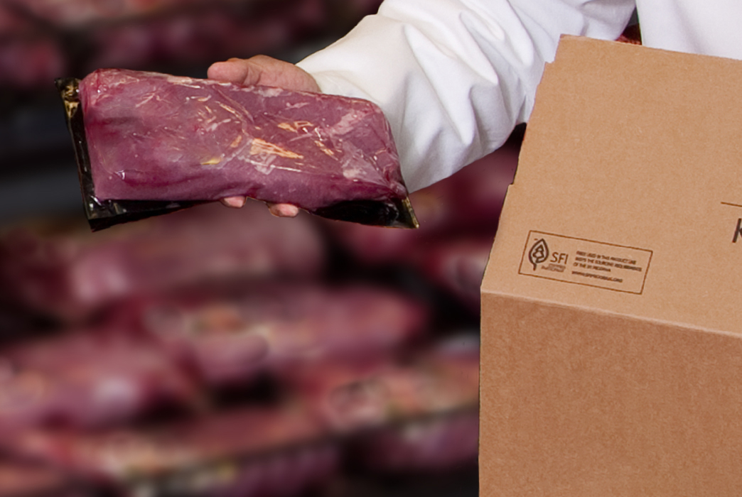 Person in a white coat holding a vacuum-sealed beef cut next to a cardboard box.