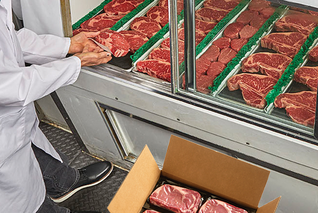 Worker stocking refrigerated display shelves with packaged beef steaks from an open box.
