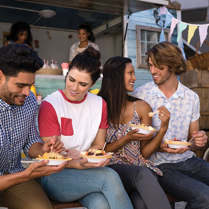 Group of young adults sitting together outdoors eating food from bowls.