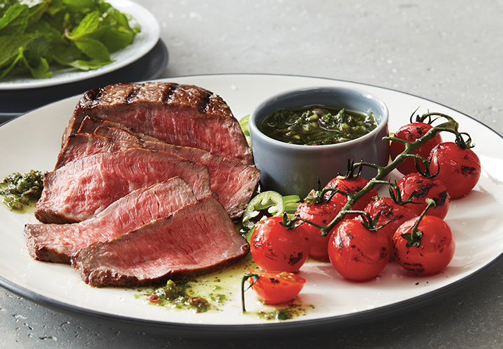 Plate of sliced medium-rare steak with grilled vine cherry tomatoes and a small bowl of green herb sauce