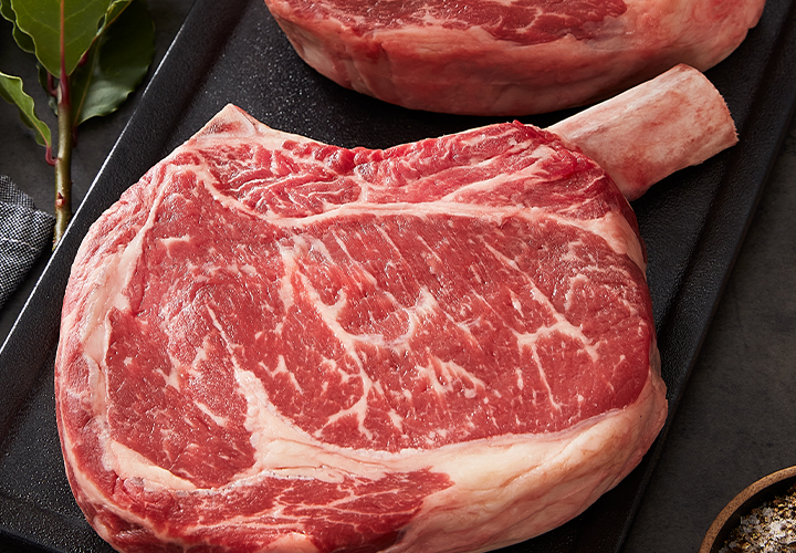 Raw tomahawk ribeye steak on a black tray with bay leaves, onions, and a dish of salt and pepper on the left