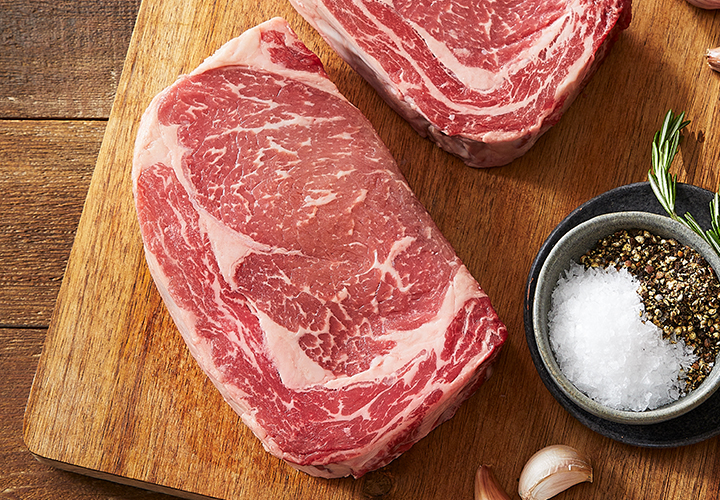 Two raw marbled ribeye steaks on a wooden board with garlic cloves, rosemary, and a bowl of salt and pepper