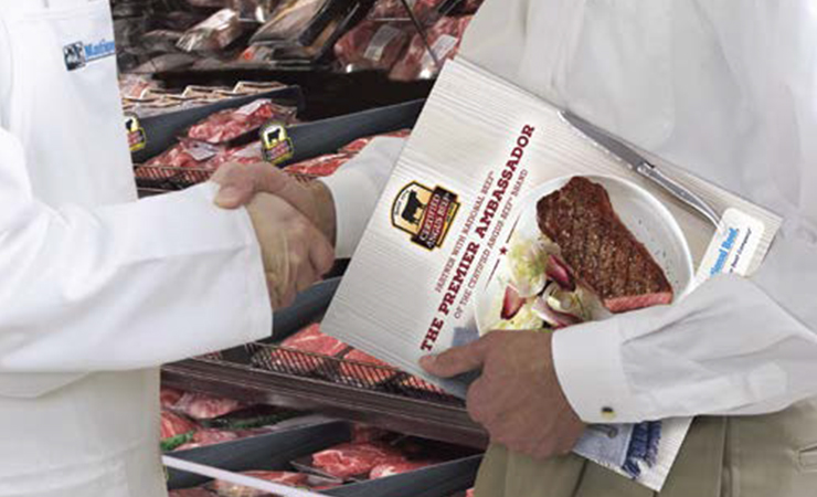Two people in white coats shake hands while holding a Certified Angus Beef brochure in front of a meat case