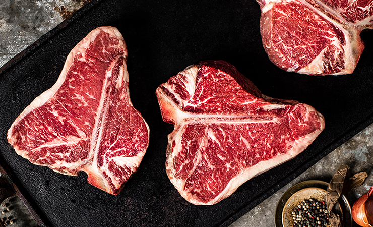 Three raw T-bone or porterhouse steaks arranged on a dark griddle with spices and aromatics nearby