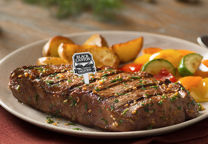 Close-up of a grilled steak with a "Black Canyon Angus Beef" label, roasted potatoes, and vegetables.