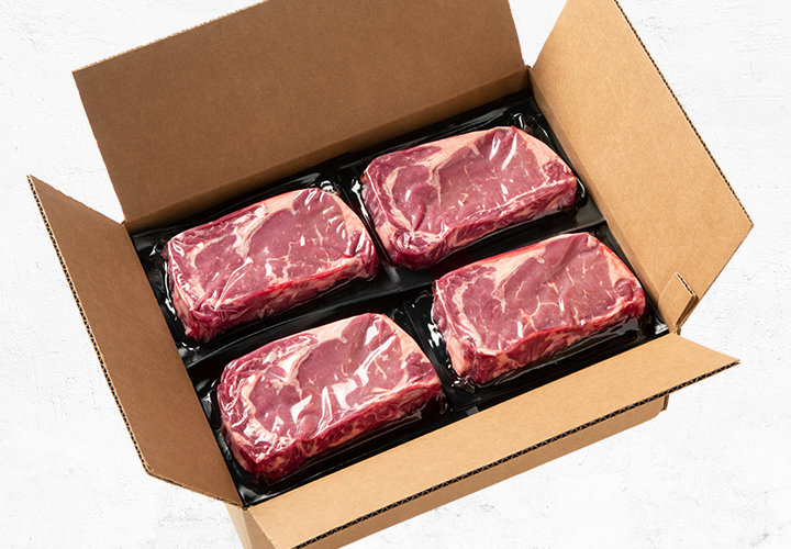 Open cardboard box containing four vacuum-sealed beef steaks.