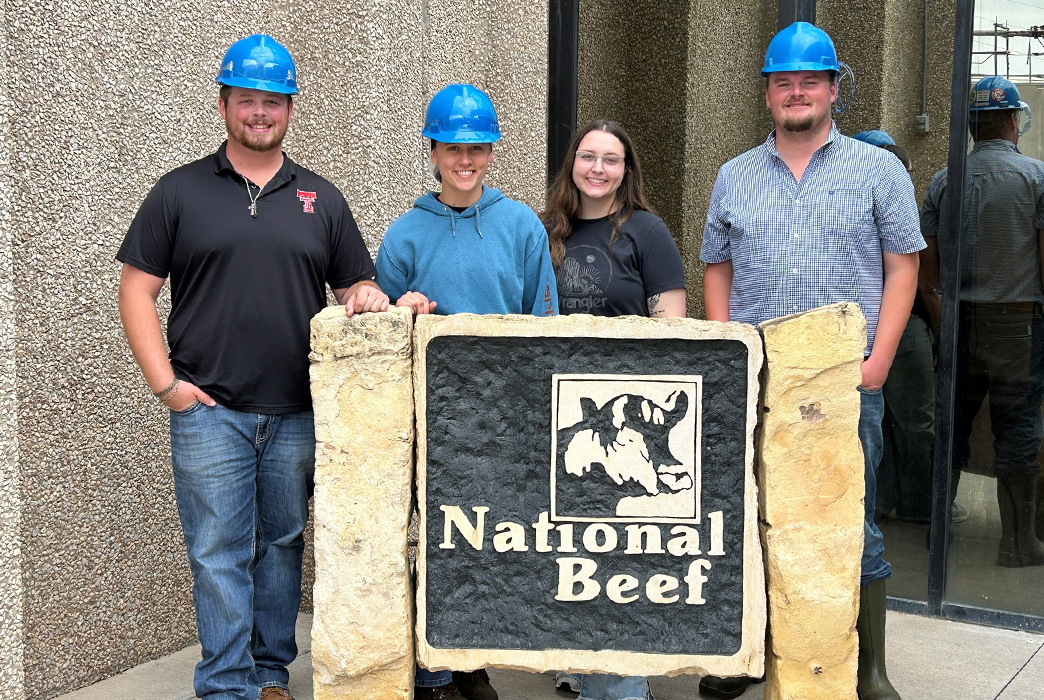 Four workers wearing blue hard hats standing by a company sign.