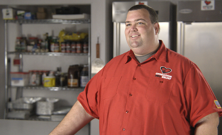 Bryan Mrocka wearing an “Operation BBQ Relief” shirt smiling in a kitchen.