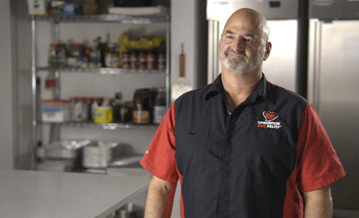 David Marks wearing a black “Operation BBQ Relief” shirt standing in a kitchen.