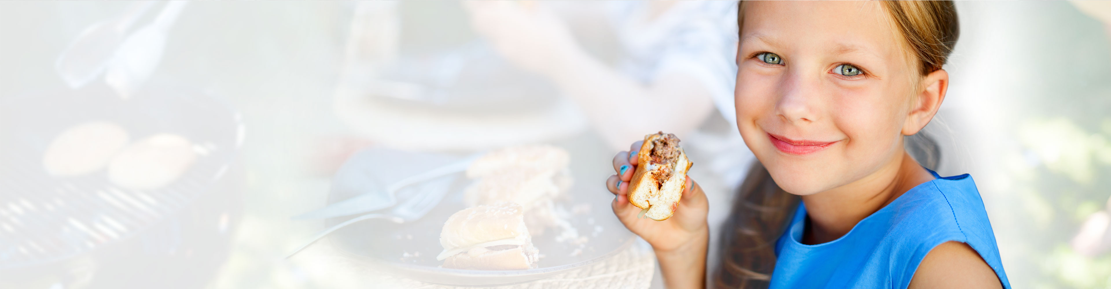 Smiling young girl holding and eating a hamburger outdoors.