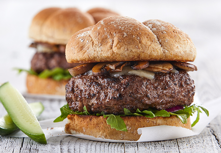Juicy beef burger with mushrooms and greens on a bun, with another burger blurred in the background.