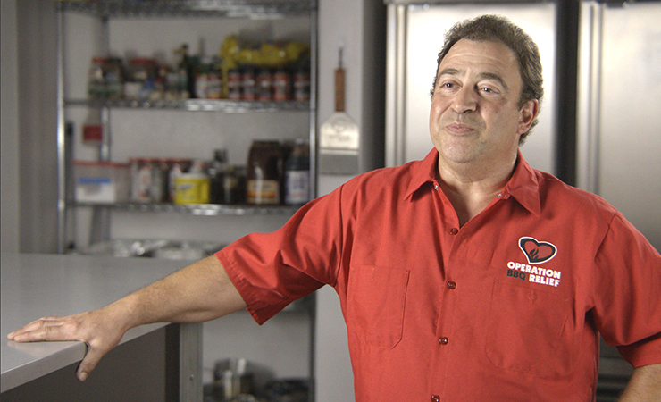 Johnny Imbrilio wearing an “Operation BBQ Relief” shirt standing in a kitchen.