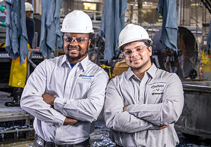 Two factory employees in white hard hats and branded shirts posing together.