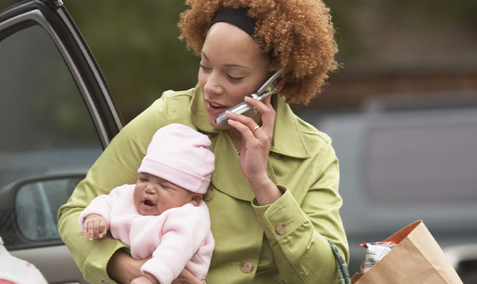 Woman holding a crying baby while talking on the phone near a car.