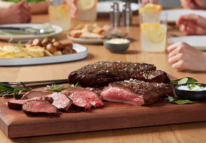 Sliced steak served on a wooden board with people dining in background.