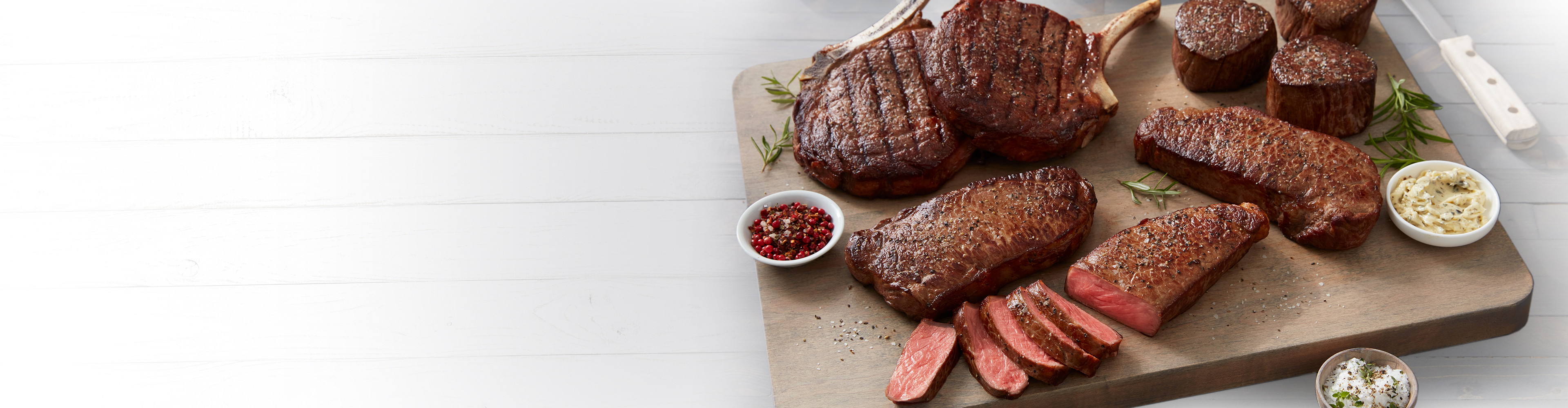 A selection of cooked steaks on a wooden board with condiments and garnishes.