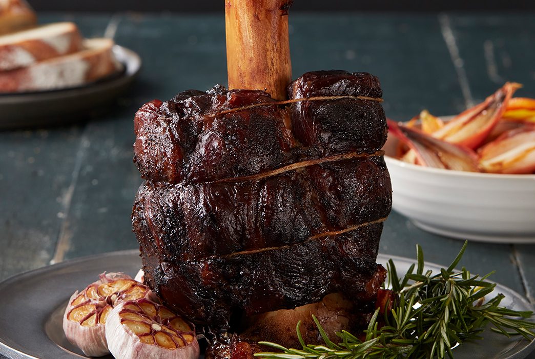 Cooked beef shank standing upright on a plate with herbs and roasted garlic.