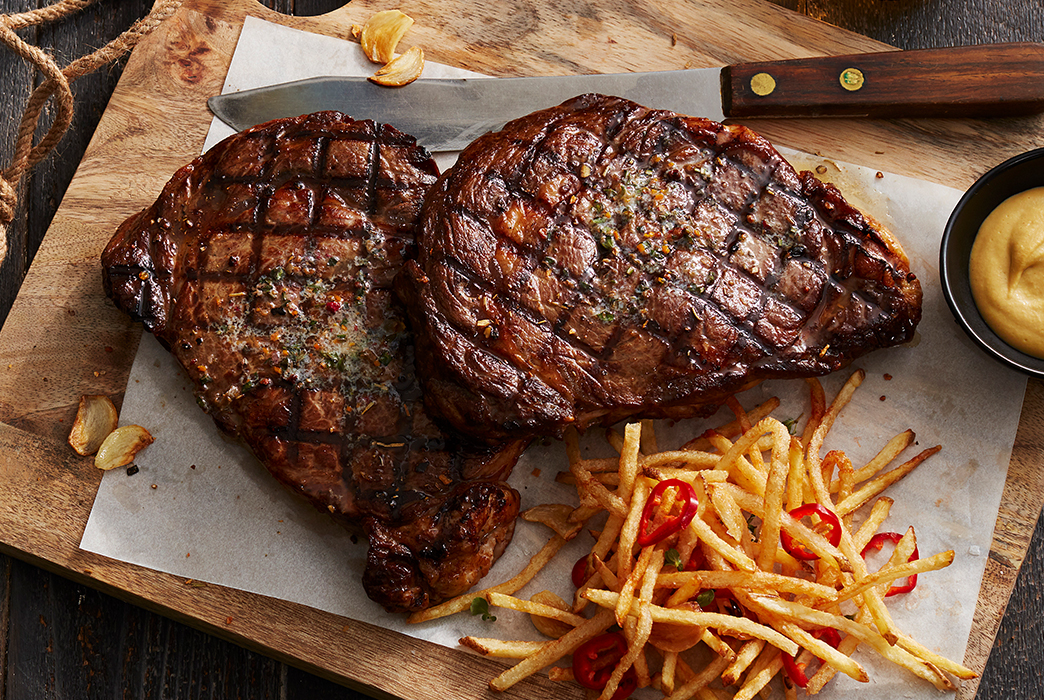 Two grilled steaks with crosshatch marks served with shoestring fries and a cup of dipping sauce on a wooden board