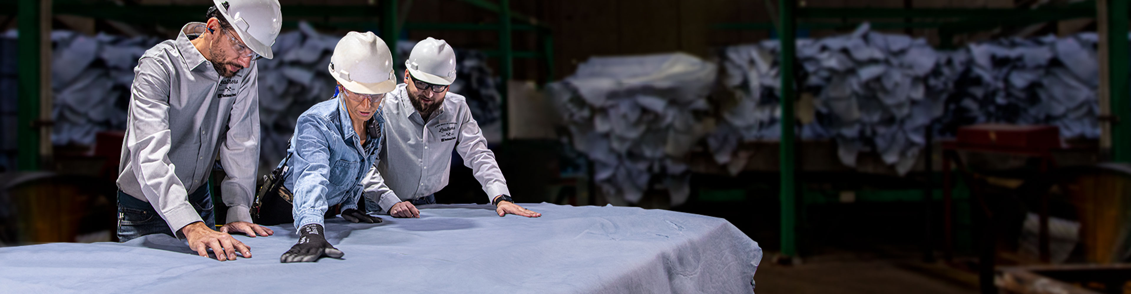 Three workers in hard hats inspecting a large sheet of leather in a factory.