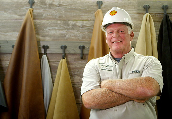 Smiling worker in a hard hat standing with arms crossed in front of hanging leather hides.
