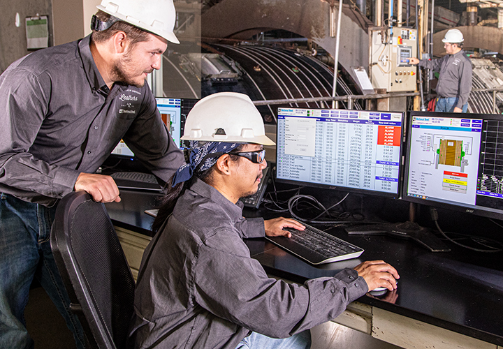 Two workers in hard hats monitoring industrial equipment on multiple computer screens.