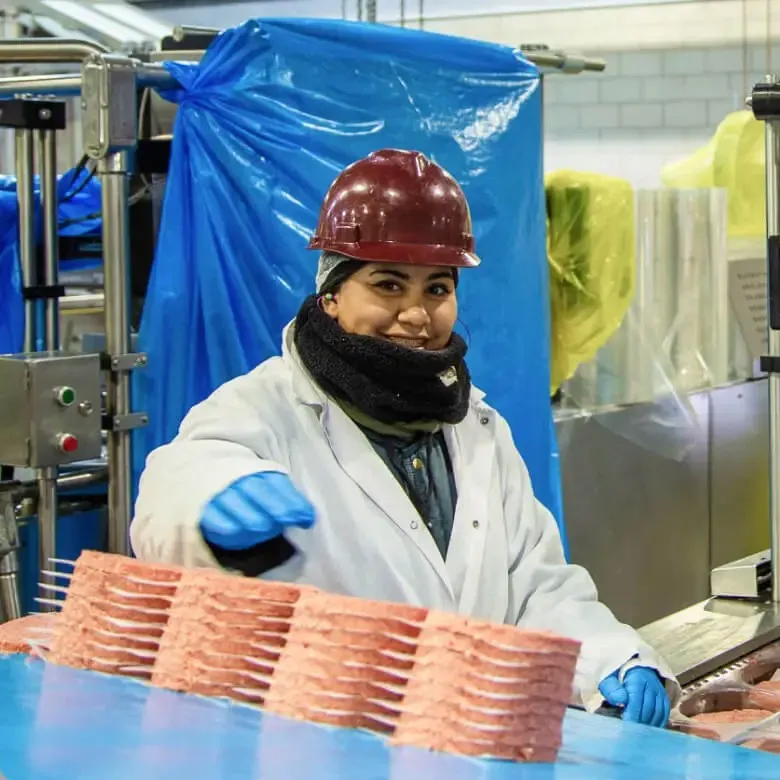 Worker in a processing facility smiling while handling stacks of formed meat patties.