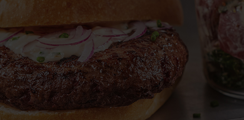 Close-up of a cooked beef burger on a bun with onions, beside a side salad on a dark background.