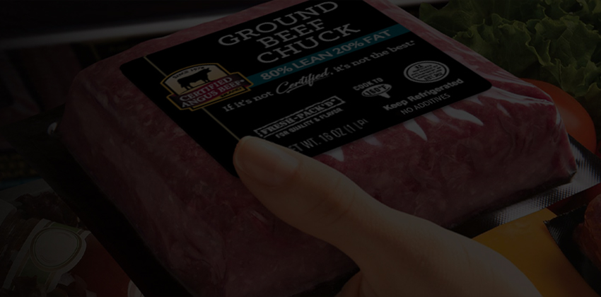 Shopper holding packaged ground beef near fresh vegetables in a grocery meat case.