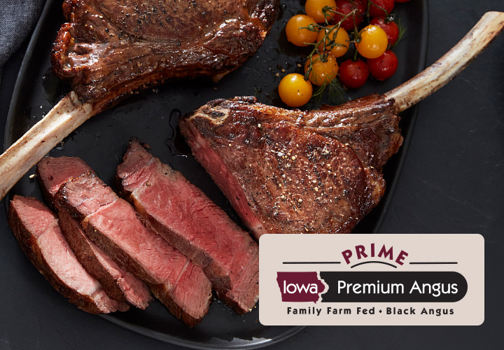 Two tom-a-hawk ribeye steaks on a dark grey plate with one partially sliced into strips and some cherry tomatoes. The plate sits on a dark grey background and the Iowa Premium Beef Prime logo is in the lower right corner partially imposed over the sliced steak.