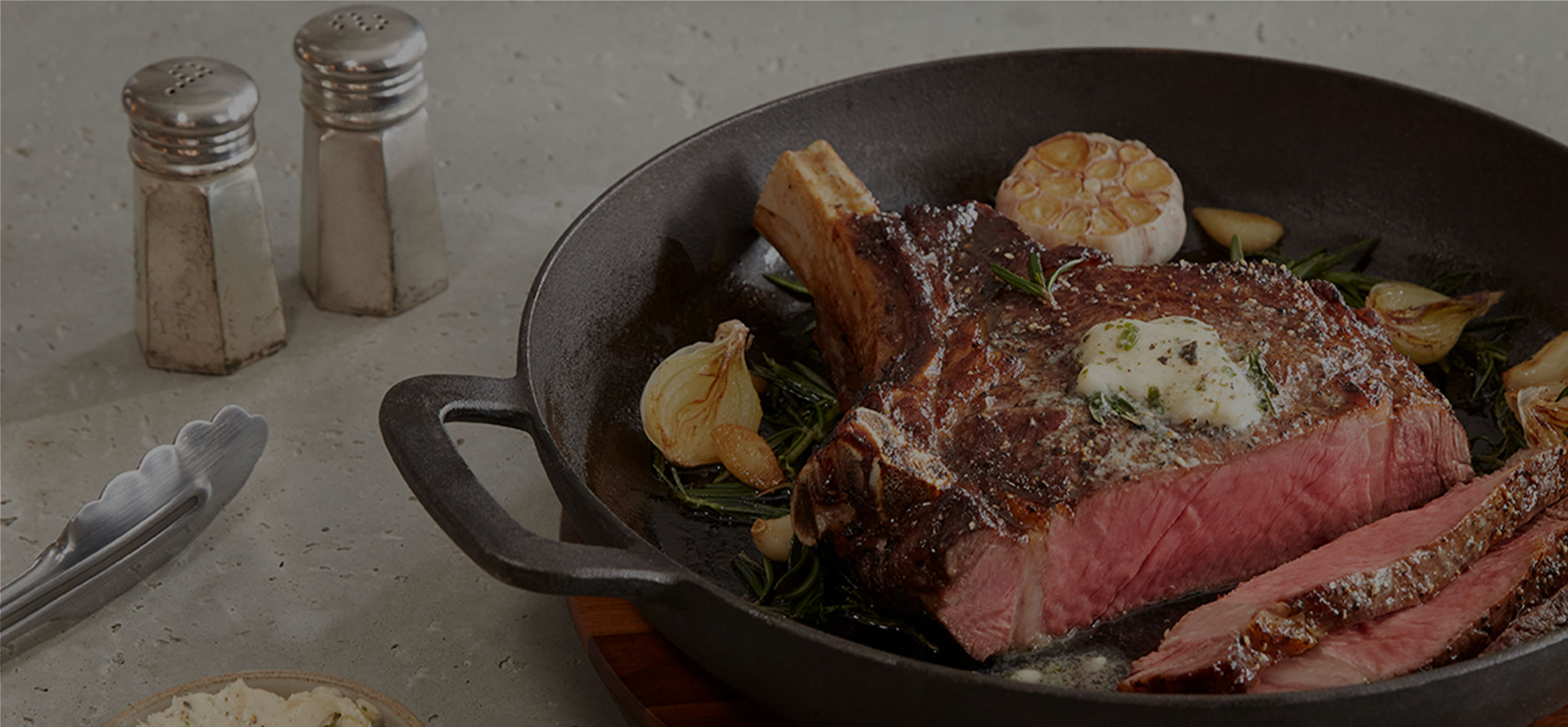 A sliced bone-in ribeye in a pan with herb butter on top, surrounded by rosemary and garlic. The pan sits upon a counter top with tongs, butter in a dish, and salt and pepper shakers next to it.