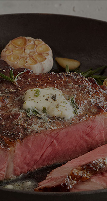 Close-up of sliced bone-in ribeye in a pan with herb butter on top and garlic behind the steak in the pan.